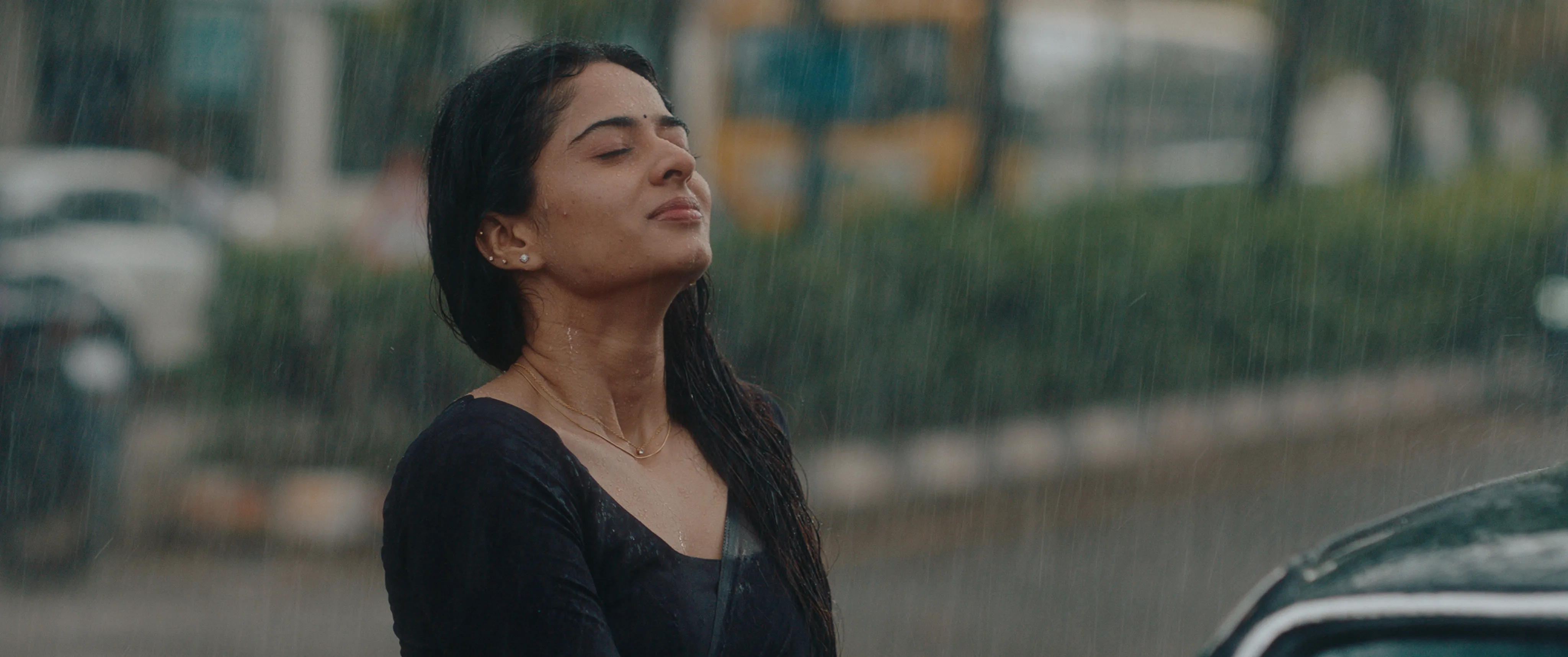 Woman in a black dress standing beside a car with her eyes closed in the rain.
