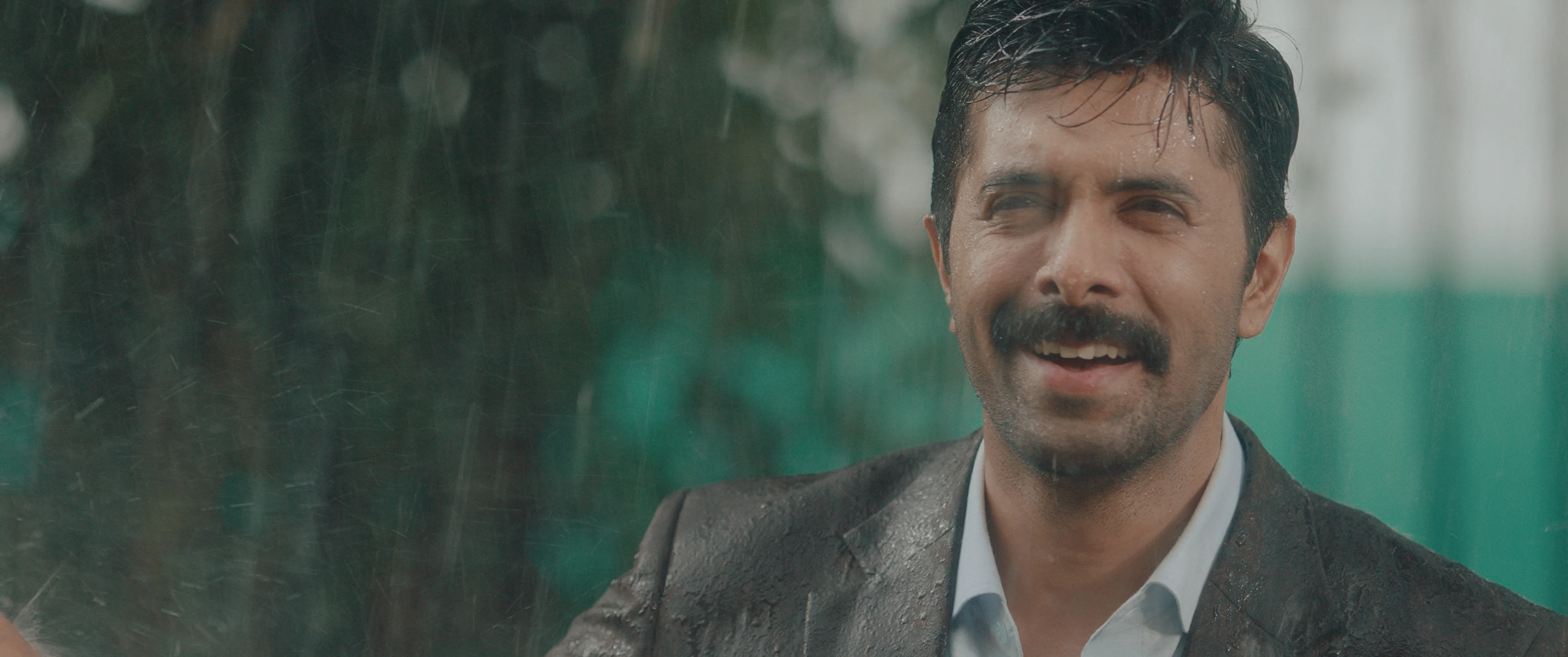 Man in a grey suit smiling while standing in heavy rain.