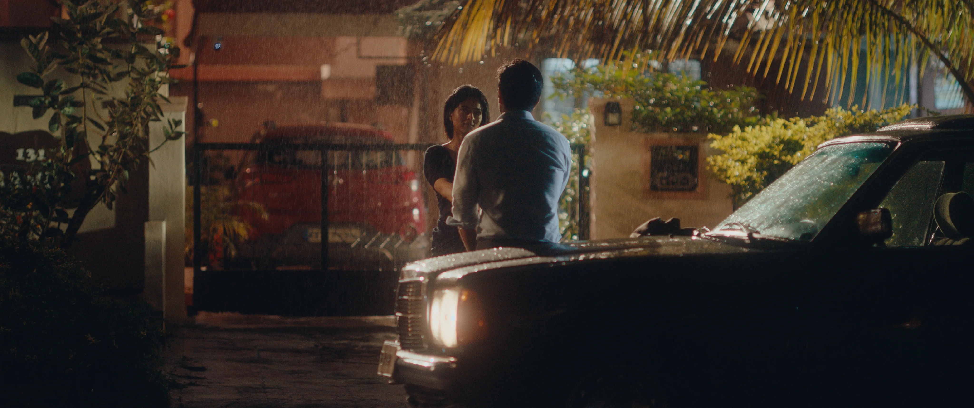 Man standing beside a black car while speaking to a woman near a gate in the rain at night.