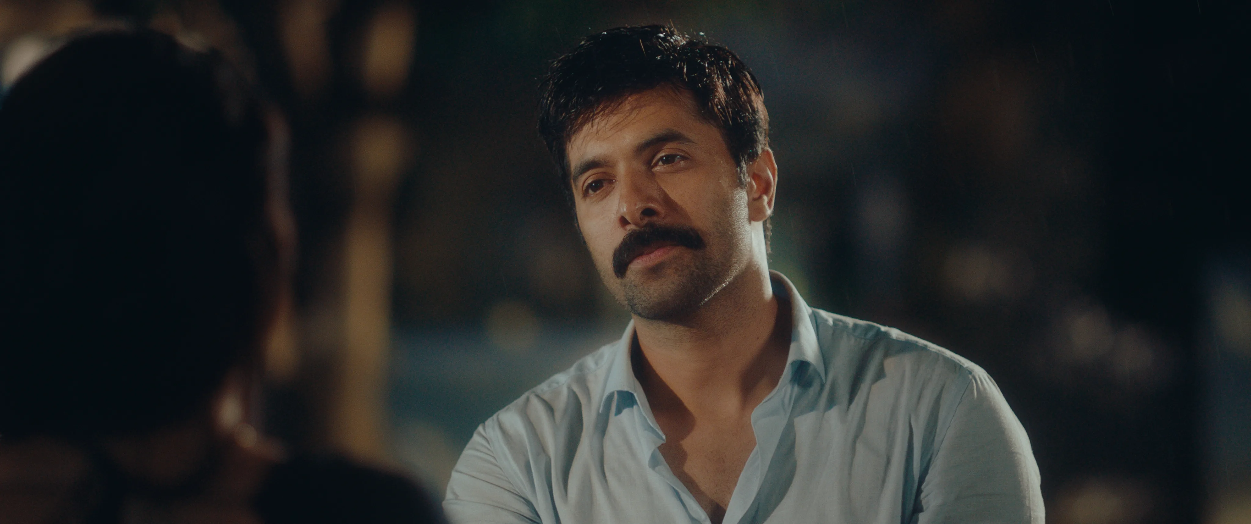 Man in a white shirt seated outdoors at night, speaking to a woman across from him.