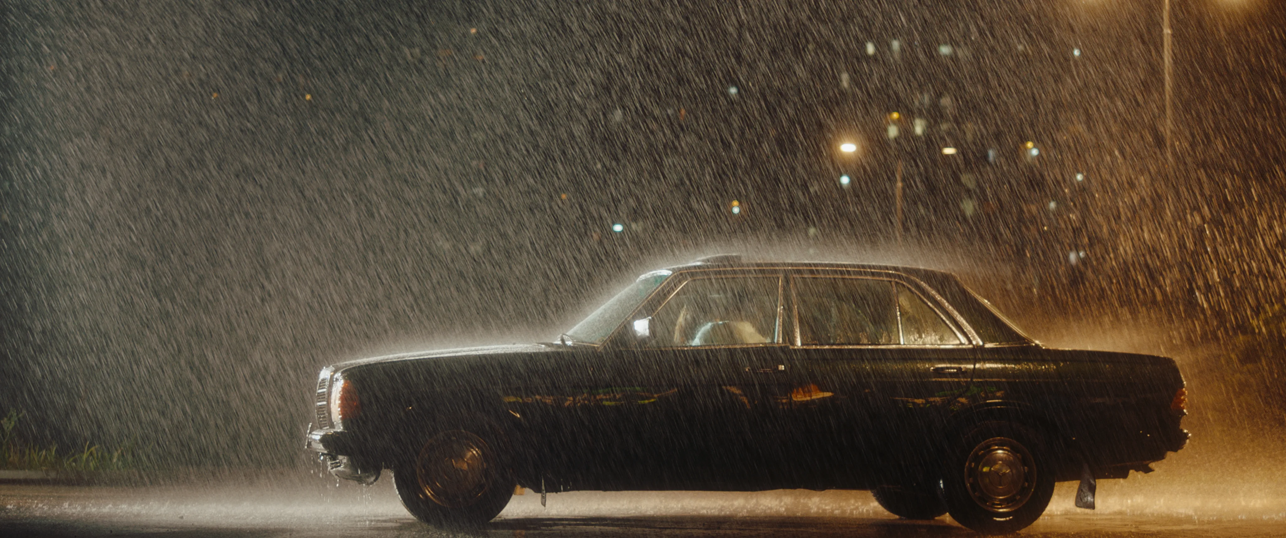 Black vintage car driving through heavy rain at night, lit by warm streetlights.