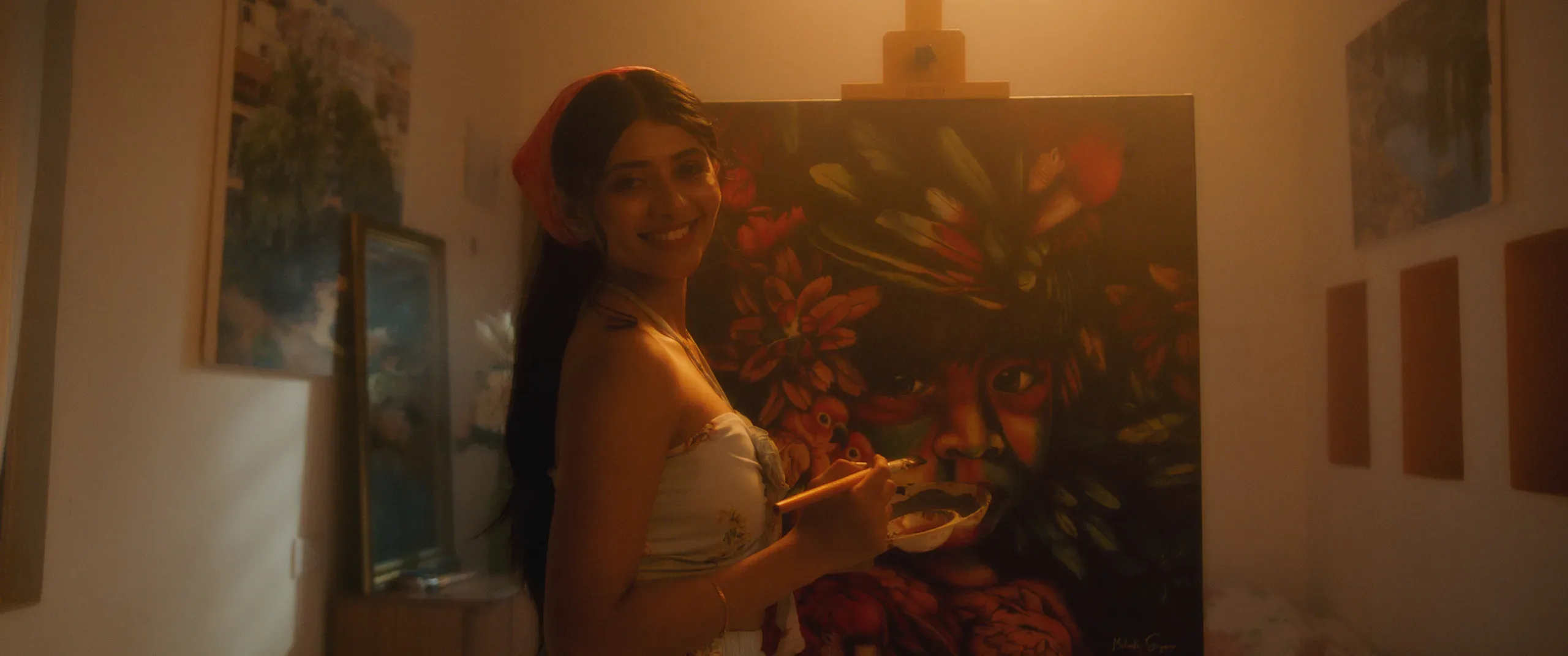 A woman in a white top holds a painter's palette beside a large, vividly colored floral painting.