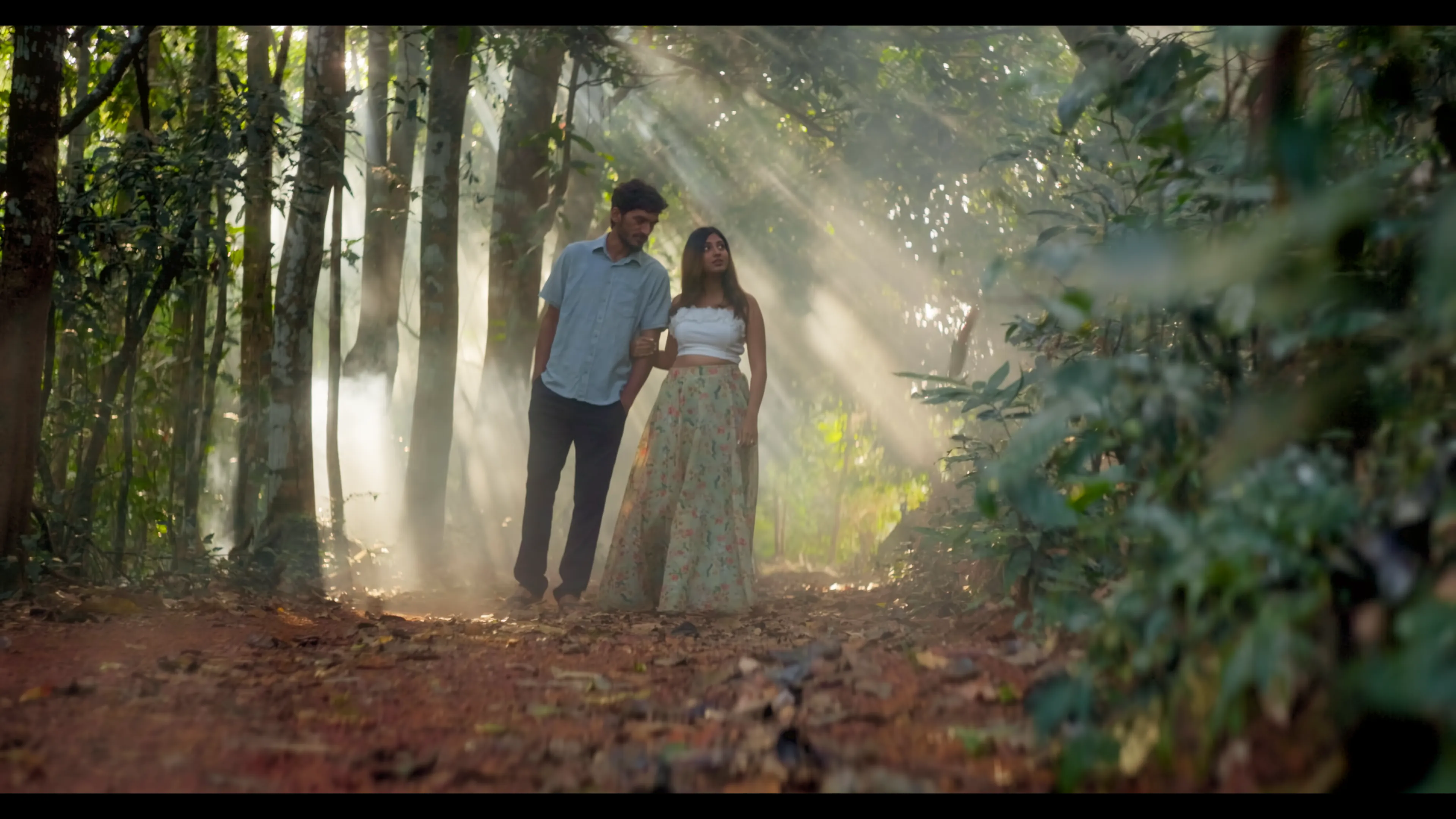 Toxench Zanv — full-frame still: a couple in a sunlit grove with light shafts through the canopy
